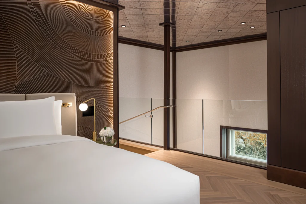 A modern hotel room or suite featuring a white bed, geometric patterned wall, wooden floor, glass railing, and a small window near the floor.